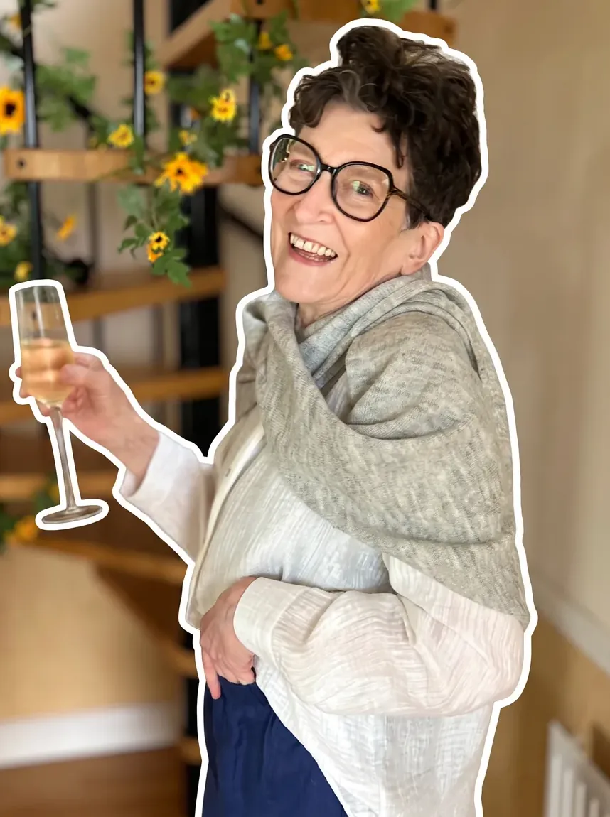 Isobel smiling with a drink in her white linen shirt, blue trousers and grey wrap